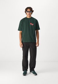 T-shirt en coton vert foncé avec un col rond et des manches courtes, arborant le texte rouge « L'homme de la nuit ». Associé à un pantalon noir et des baskets vertes.