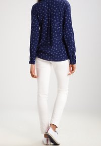 Camicia a maniche lunghe blu navy con piccoli motivi bianchi, abbinata a pantaloni bianchi slim-fit e sneakers bianche con dettagli colorati.