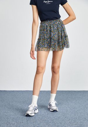 Femme portant un t-shirt bleu marine, une jupe courte fleurie, des chaussettes blanches et des chaussures de course blanches, debout sur un sol recouvert d'un tapis bleu.