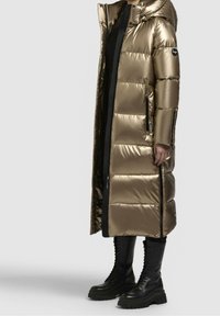 Metallisch goldene Pufferjacke mit Kapuze, langer Schnitt, mit Reißverschlusstaschen und horizontaler Steppung. Kombiniert mit schwarzen Schnürschuhen im Combat-Stil.