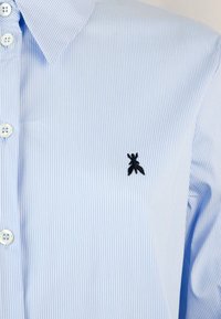 Camicia a strisce blu chiaro con logo ricamato nero sul petto, dotata di colletto classico e chiusura con bottoni.