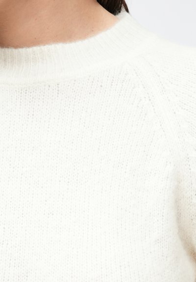 Λευκό πλεκτό πουλόβερ με ριμπ λαιμόκοψη και υφασμάτινη υφή. Κοντινή λήψη αναδεικνύει το μαλακό, αέρινο υλικό και το διακριτικό μοτίβο.