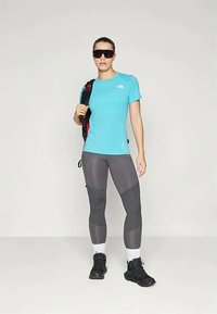 Γυναίκα που φορά μαύρες ορειβατικές μπότες, γκρι κολάν, τυρκουάζ κοντομάνικο μπλουζάκι, κρατάει μαύρο σακίδιο και μεγάλα σκούρα γυαλιά ηλίου, στέκεται σε λευκό πάτωμα.