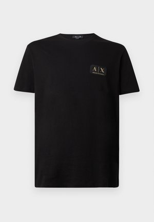 Camiseta de algodón negra con mangas cortas y cuello redondo. Presenta un pequeño parche de logo en negro y dorado en el área del pecho izquierdo.