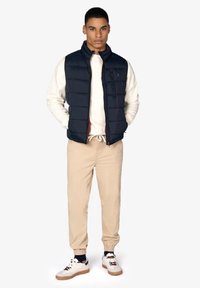 Chaleco de plumas navy sobre un suéter ligero, pantalones beige ajustados y zapatillas blancas con detalles en marrón. Atuendo casual en capas con cierre visible.