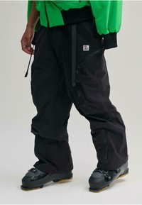 Pantaloni impermeabili neri presentano multiple tasche con cerniera, cinturini regolabili e una vestibilità ampia, abbinati a scarponi da sci neri con un design robusto.