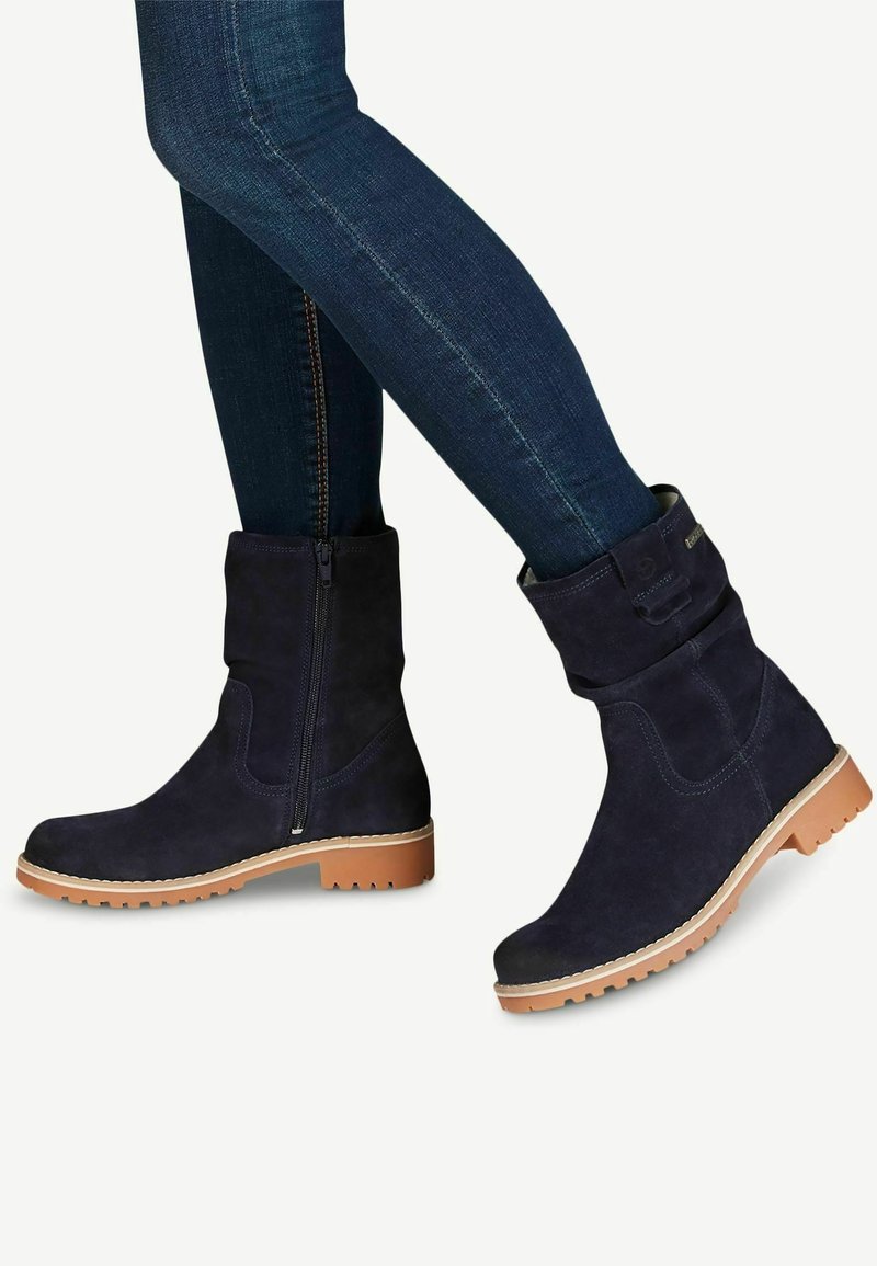 Bottines en suede bleu marine foncé avec une fermeture éclair sur le côté, présentant une tige texturée, une semelle en caoutchouc beige clair contrastante et des détails de couture minimaux.