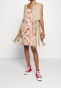 Kvinna i en blommig klänning med en beige fransväst och röda högstövlar, som står mot en vit bakgrund.