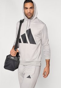 Ανοιχτό γκρι φούτερ με κουκούλα και μαύρο λογότυπο, κορδόνι στην κουκούλα, ραβδωτές μανσέτες. Το μοντέλο κρατάει μια μαύρη, υφασμάτινη τσάντα γυμναστηρίου με ιμάντα ώμου.