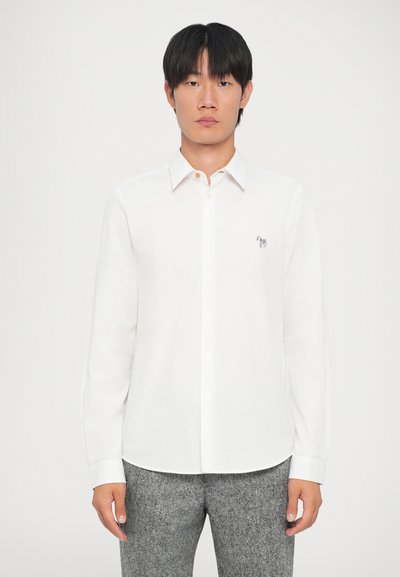Camicia bianca a maniche lunghe con colletto, apertura frontale con bottoni e dettaglio ricamato sul petto. Tessuto testurizzato abbinato a pantaloni grigi.