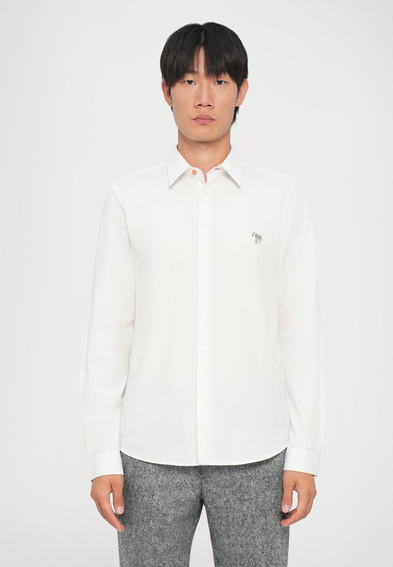 Camicia bianca a maniche lunghe con colletto, apertura frontale con bottoni e dettaglio ricamato sul petto. Tessuto testurizzato abbinato a pantaloni grigi.