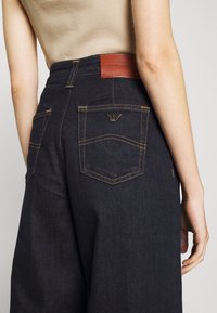 Mörka denimbyxor med vida ben och hög midja, med kontrasterande sömmar, två bakfickor och en brun läderlogotyp.