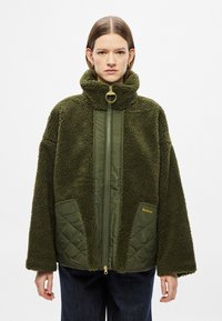 Olivekrohvitud roheline üle suurusega jakk pehme sametise kangaga, millel on kõrge krae, kuldsed detailid ja tikandiga taskuaktsendid.