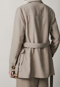 Beżowy trencz z paskiem, długimi rękawami, bocznymi kieszeniami i wentylowanym tyłem. Posiada klasyczny kołnierz i gładką fakturę.