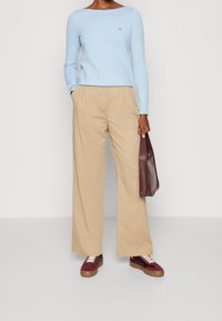 Top azzurro chiaro a maniche lunghe, pantaloni beige a gamba larga, sneakers bordeaux e crema, e una borsa bordeaux. Materiale in cotone, design casual.