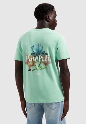 Man die wegdraait, draagt een muntgroen T-shirt met een bloemmotief en de tekst "Pure Path" op de rug, gecombineerd met lichtblauwe spijkerbroek.
