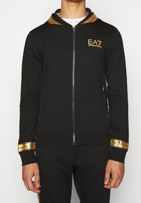 Svart zip-hoodie med en guldlogo, med ett mönstrat foder i huvan och detaljer på muddar, tillverkad av ett glatt tyg.