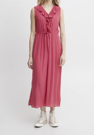 Femme portant une robe midi rose sans manches avec un col en V à volants et une ceinture nouée, associée à des baskets montantes blanches.