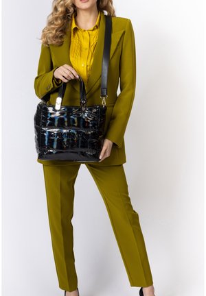 Frau in olivgrünem Anzug hält schwarze glänzende Gattinoni Roma Handtasche mit doppelten Griffen und Schulterriemen vor weißem Hintergrund.