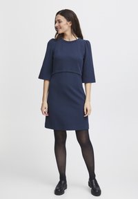 Marinblå klänning med trekvartsärmar, texturerad tyg, figurnära midja och knälång längd. Bärs med svarta strumpbyxor och loafers.