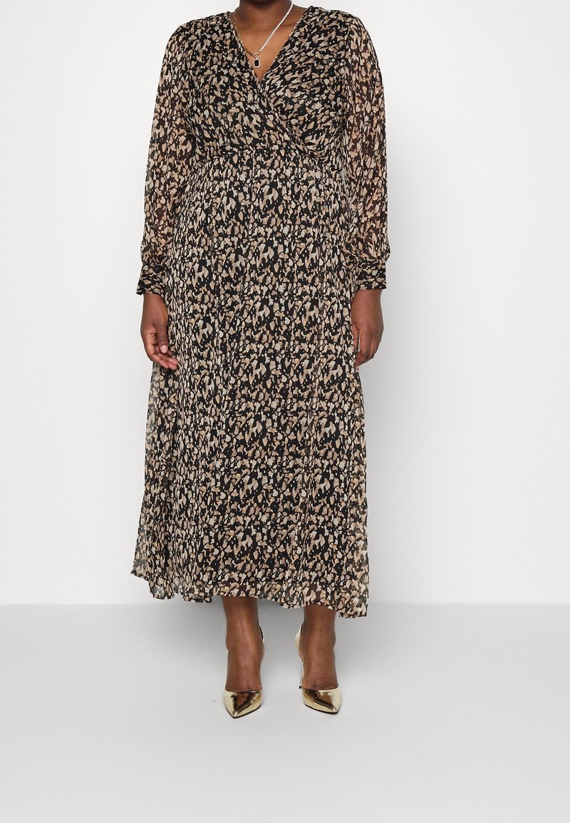 Femme portant une robe à manches longues, longue jusqu’aux chevilles, noire et beige à motifs, des escarpins dorés à bouts pointus et un collier avec pendentif.