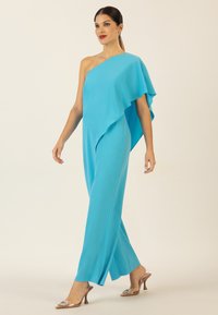 Un jumpsuit lungo fino al pavimento, con una spalla sola, di un vibrante color turchese, caratterizzato da un design asimmetrico drappeggiato e una fessura laterale, abbinato a tacchi trasparenti.