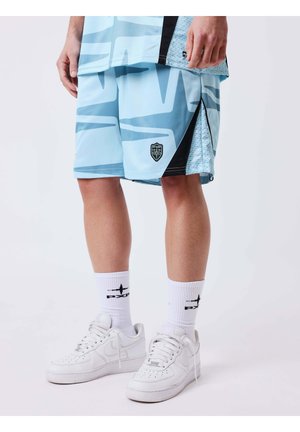 Personne portant un short de sport bleu clair à motifs, des chaussettes blanches mi-mollet avec un logo noir, et des baskets blanches, debout sur un sol blanc.