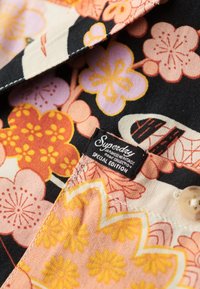 Camisa con cuello que presenta un patrón floral en tonos rosa, naranja y amarillo sobre un fondo negro. Incluye una etiqueta que dice "Superdry Edición Especial".