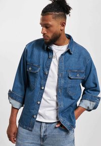 Lyseblå denimskjorte med knapper foran, to brystlommer, rullede ermer og kontrasterende lysere mansjetter, kombinert med en hvit t-skjorte under.