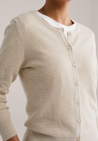 Béžový pletený kardigán s textúrovaným vzorom, okrúhlym výstrihom, zapínaním na gombíky a rebrovanými manžetami. Jednoduchý a priliehavý dizajn.
