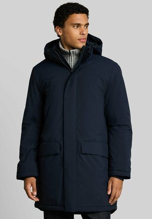 Man draagt een donkerblauwe winterjas met capuchon en een grijze gebreide trui, staat en kijkt naar opzij tegen een effen achtergrond.