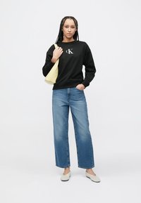 Czarna bluza z kapturem z logo "CK", jasnoniebieskie dżinsy o szerokich nogawkach, żółta torebka na ramię oraz jasnoszare buty wsuwane. Gładki materiał i luźny krój.