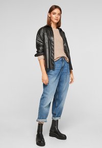 Zwarte leren overhemdjasje over een beige gebreide trui, gecombineerd met lichtblauwe high-waisted jeans en zwarte enkellaarzen. Oprol-detail op de jeans.