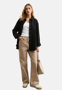 Zwarte, knoopsluiting shirt met textuur, beige wijde broek, beige handtas en witte schoenen. Model staat met handen op de heupen.