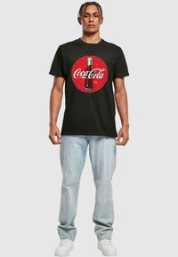 Sort bomulds T-shirt med et stort retro Coca-Cola flaske grafik på en rød cirkel, parret med lyseblå jeans og hvide sneakers.