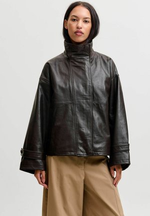Femme aux cheveux noirs et raides portant une veste en cuir noire à col montant et un pantalon beige ample, debout devant un fond uni.