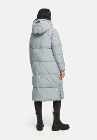 Lang, lysegrå pufferfrakke med hætte, der har lodrette syninger og knaplukninger i siden. Båret over sorte bukser og støvler.