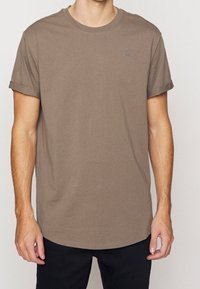 T-shirt court à manches courtes marron foncé avec un col rond, des manches légèrement retroussées et un petit logo sur la poitrine. Texture du tissu en coton doux.