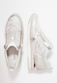 Białe sneakersy z metalicznymi srebrnymi akcentami, fakturowaną cholewką w różnych odcieniach, zapinane na suwak po boku oraz gumową podeszwą z lekkim podwyższeniem.