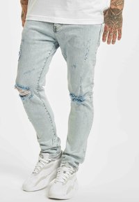 Mies, który ma na sobie jasnoniebieskie poszarpane skinny jeans i białe sneakersy, stoi z jedną widoczną ręką, na której widać tatuaż sowy i liczby na palcach.