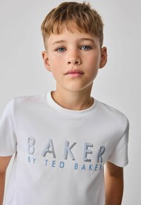 Bela bomba z bombažem z napisom "BAKER" v modrih in črnih črtastih črkah, z "by TED BAKER" v manjšem tisku spodaj. Kratke rokave, okroglo izreza.