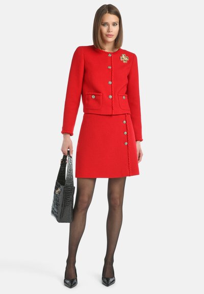 Giacca da tailleur rossa con texture, abbinata a una gonna, con bottoni e tasche. La modella tiene una borsa a tote nera con stampa cocco.