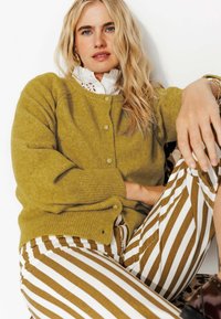 Cardigan vert olive avec texture côtelée et fermeture à boutons, associé à un pantalon rayé vertical marron et blanc.