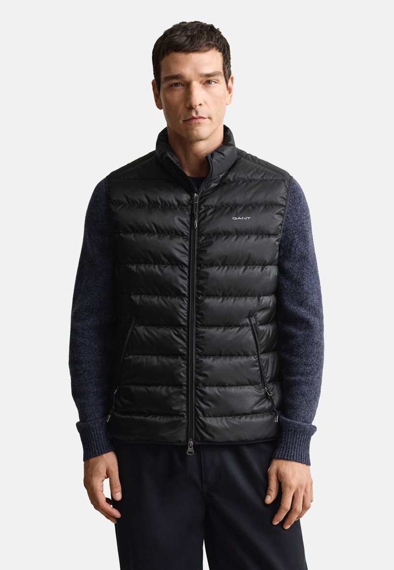 Schwarze gesteppte Weste mit hohem Kragen, Reißverschluss und Seitentaschen; getragen über einem dunkelblauen strukturierten Pullover. Minimalistisches Branding auf der Brust.