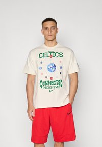 Kremfarget t-skjorte med "Boston Celtics" grafikk i grønt, med en jordklode og basketballer. Kombinert med røde Nike-shorts.
