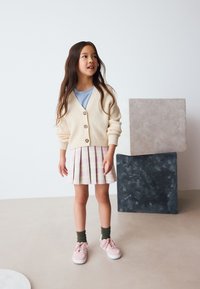 Cárdigan de punto color crema con botones, camiseta azul claro, falda plisada de cuadros rosa, calcetines verdes y zapatillas rosas; sobre un fondo de bloques geométricos.