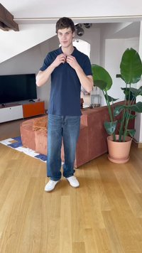 Jūros laivyno polo marškiniai, džinsai ir balti sportbačiai. Vidaus aplinka su dideliu augalu ir oranžiniais akcentais apstatyta sofa fone.