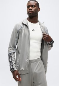 Άνδρας που φορά γκρι ζακέτα με φερμουάρ και ταιριαστό παντελόνι, κρατώντας τη ζακέτα ανοιχτή για να αποκαλύψει ένα λευκό πουκάμισο με μαύρο λογότυπο στο στήθος.