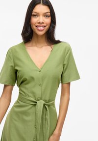 Femme souriante, portant une robe verte à manches courtes avec des boutons et une ceinture nouée, sur fond blanc uni.