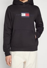 Black hoodie made of cotton blend, features a front pocket, ribbed cuffs, and a logo design in red, white, and blue across the chest.